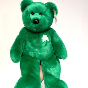 May include: A plush green teddy bear with a white shamrock on its chest. The bear has a black nose and a tag attached to its ear. The bear is a vibrant shade of green and has a soft, furry texture.