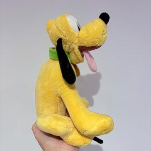 May include: A yellow plush toy dog with black ears, a black nose, and a pink tongue. The dog is wearing a green collar. The toy is held in a hand.