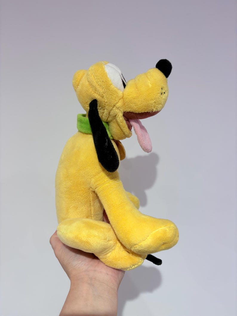 May include: A yellow plush toy dog with black ears, a black nose, and a pink tongue. The dog is wearing a green collar. The toy is held in a hand.
