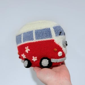 May include: A knitted toy bus in red, white, and blue. The bus features a white roof, blue windows, and red body with white flower accents. The wheels are black and gray. The toy is held in a hand.