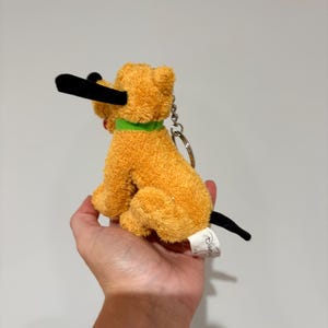 Puede incluir: Un llavero de peluche de perro naranja con nariz y orejas negras y un collar verde. El perro est&aacute; sentado, con una etiqueta adjunta. Producto Disney.