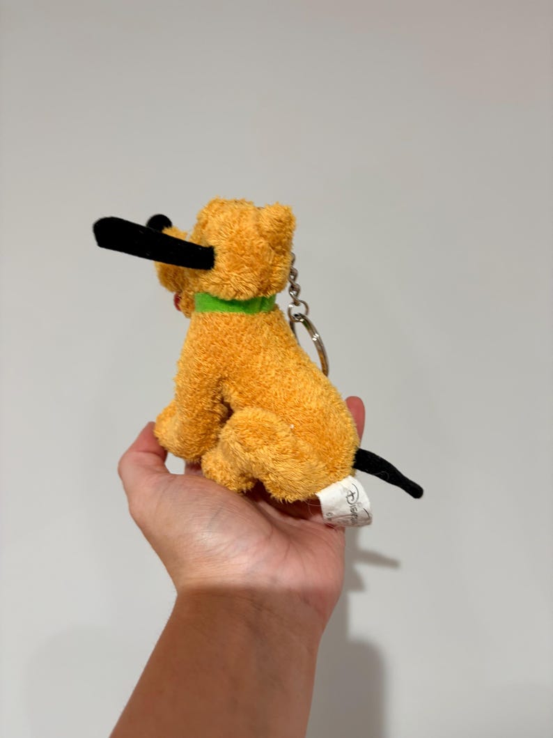 Puede incluir: Un llavero de peluche de perro naranja con nariz y orejas negras y un collar verde. El perro est&aacute; sentado, con una etiqueta adjunta. Producto Disney.