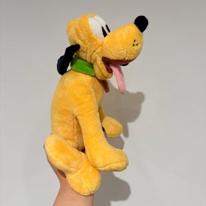 May include: A plush toy of Pluto, a yellow dog with black ears, a green collar, and a pink tongue sticking out. The toy is held up against a white background.