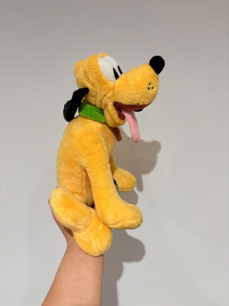 May include: A plush toy of Pluto, a yellow dog with black ears, a green collar, and a pink tongue sticking out. The toy is held up against a white background.