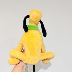 May include: A yellow plush toy dog with black ears and tail, and a green collar. The dog is sitting with its legs spread out. The toy is a popular character from a cartoon.