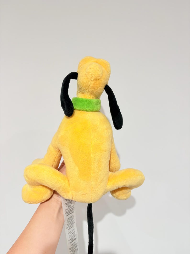 May include: A yellow plush toy dog with black ears and tail, and a green collar. The dog is sitting with its legs spread out. The toy is a popular character from a cartoon.
