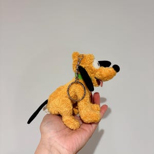 Puede incluir: Un peluche de Pluto, un perro amarillo con orejas negras y una cola negra. El juguete tiene un collar verde y un llavero de metal. La lengua del perro est&aacute; fuera. El juguete se sostiene en una mano.