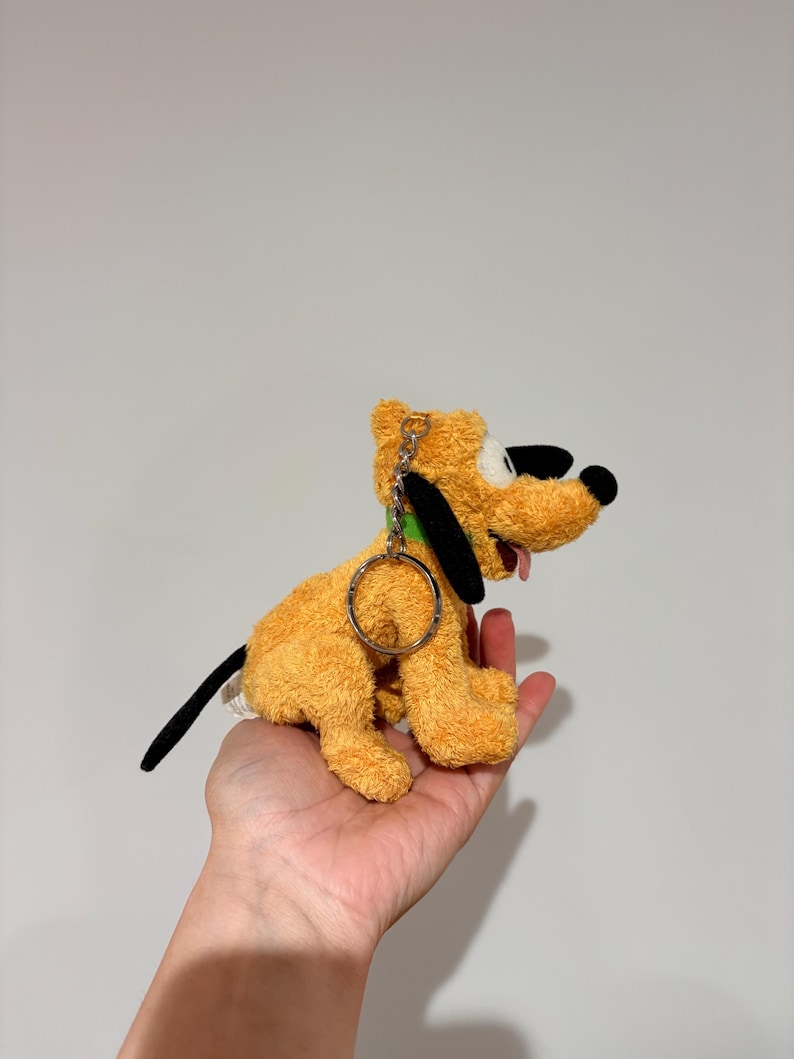 Puede incluir: Un peluche de Pluto, un perro amarillo con orejas negras y una cola negra. El juguete tiene un collar verde y un llavero de metal. La lengua del perro est&aacute; fuera. El juguete se sostiene en una mano.