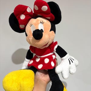 May include: A plush Minnie Mouse doll featuring a red dress with white polka dots, a matching bow, and yellow shoes. The doll has black ears, arms, and legs, and white gloves. The doll is a classic Disney character.