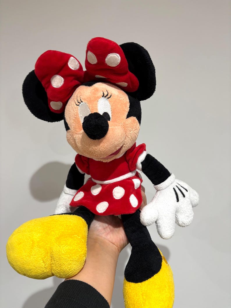 May include: A plush Minnie Mouse doll featuring a red dress with white polka dots, a matching bow, and yellow shoes. The doll has black ears, arms, and legs, and white gloves. The doll is a classic Disney character.
