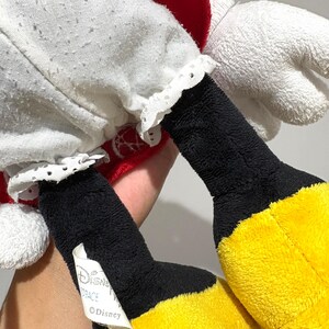 May include: Close-up of a plush Minnie Mouse doll. The doll features a white dress with a red polka-dot bow and matching red accents. The doll has black legs and yellow feet. A tag with the Disney logo is visible.