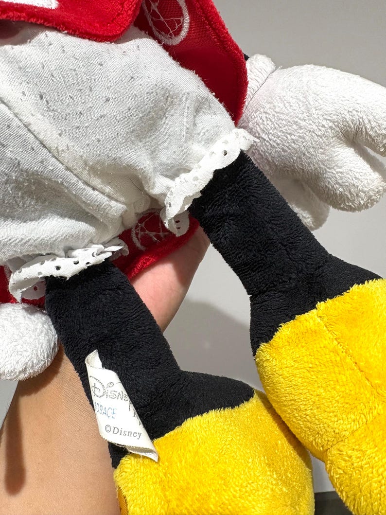 May include: Close-up of a plush Minnie Mouse doll. The doll features a white dress with a red polka-dot bow and matching red accents. The doll has black legs and yellow feet. A tag with the Disney logo is visible.