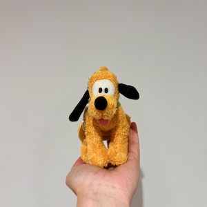 Puede incluir: Un peluche amarillo que representa un perro con orejas negras y un collar verde. El perro tiene una nariz negra y est&aacute; sentado erguido. El juguete se sostiene en la mano de una persona. El juguete es un personaje de dibujos animados popular.