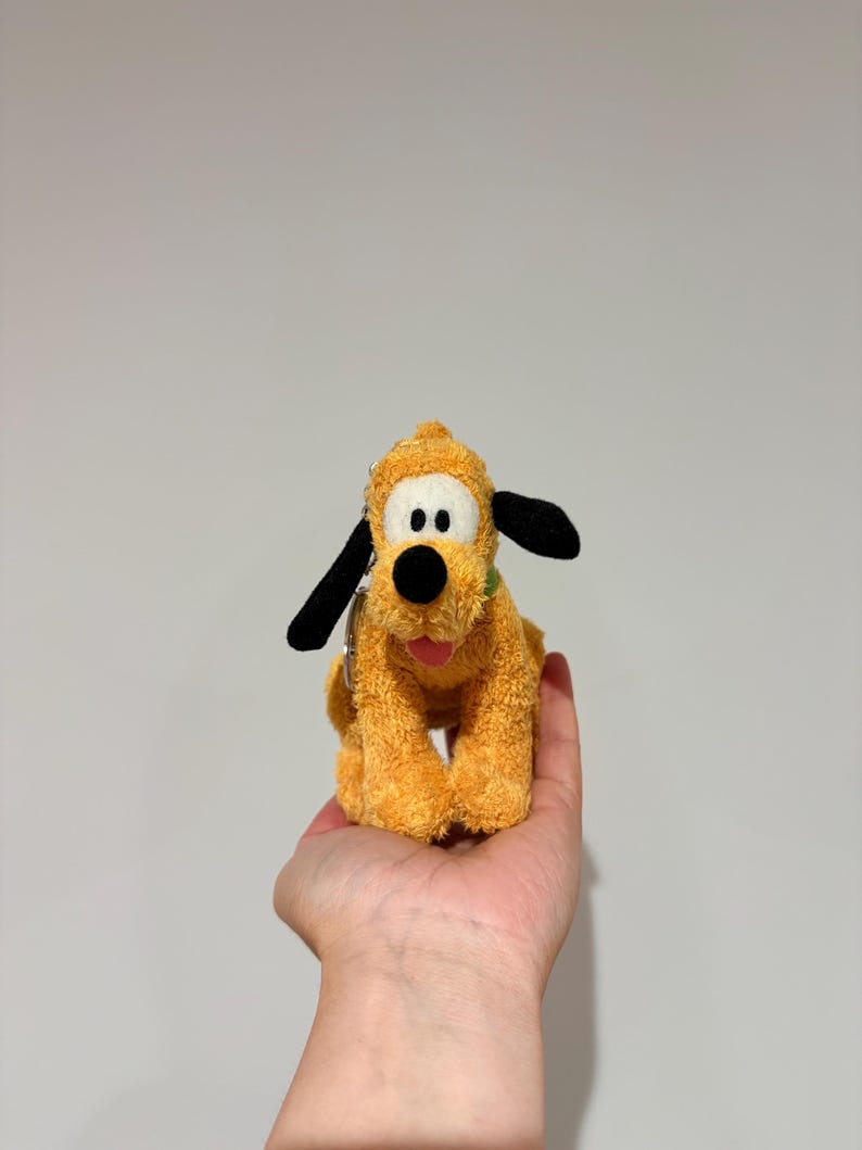 Puede incluir: Un peluche amarillo que representa un perro con orejas negras y un collar verde. El perro tiene una nariz negra y est&aacute; sentado erguido. El juguete se sostiene en la mano de una persona. El juguete es un personaje de dibujos animados popular.