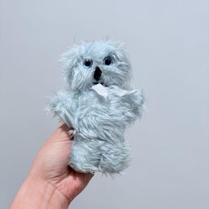 May include: A light blue plush koala puppet with large blue eyes and a black nose. The koala has a white fabric around its neck and is made of soft, fuzzy material. The puppet is held in a hand.