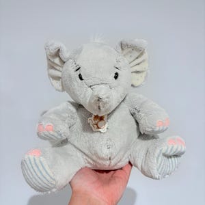 May include: A soft grey elephant toy with large ears, small black eyes, and a trunk. The elephant has pink and blue accents on its feet and ears. A small tag with a squirrel design hangs from its neck.