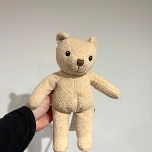 May include: A plush teddy bear in a light tan color. The bear has a textured, fuzzy exterior, small black eyes, and a brown nose. It has a simple, classic design with outstretched arms and a tag attached to its leg.