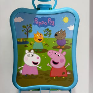 May include: A blue Peppa Pig themed carry case with a handle. The case features a colourful illustration of Peppa Pig and friends in a grassy field with a sun and trees. The case is held by a hand.