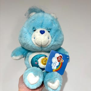 May include: A light blue Care Bear plush toy with a white belly and heart-shaped feet. The bear has a smiling face, a blue heart on its nose, and a crescent moon and star design on its belly. A tag with a rainbow and the words "Bedtime Bear" is attached.