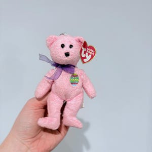May include: A pink plush bear toy with a purple ribbon and an Easter egg design. The bear has a red heart-shaped tag that reads "Basket Beanies".