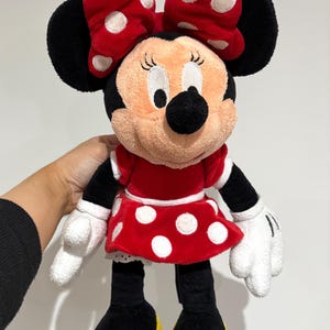 May include: A Minnie Mouse plush doll, featuring a red dress with white polka dots, a matching bow, and black ears. The doll has white gloves, black arms and legs, and yellow shoes. The plush toy is soft and cuddly.