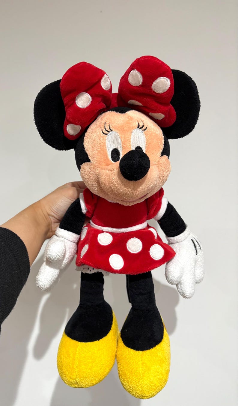 May include: A Minnie Mouse plush doll, featuring a red dress with white polka dots, a matching bow, and black ears. The doll has white gloves, black arms and legs, and yellow shoes. The plush toy is soft and cuddly.