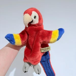 May include: A plush parrot toy in vibrant red, yellow, and blue. The toy has a white beak, gray feet, and a tag that reads "Ravensden Collection". The parrot's wings are spread, showcasing the colorful feathers.