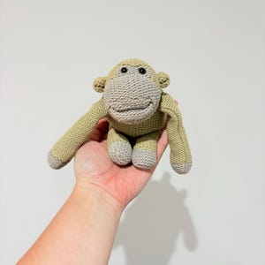 May include: A small, hand-knitted monkey toy featuring a light olive green body and limbs with contrasting light grey accents on the face, hands, and feet. The plush toy has two small black bead eyes and a stitched mouth, with a textured knit finish.