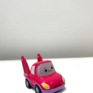 May include: A magenta toy car with a smiling face, white windows, and two upright pink fins. The car has yellow headlights, purple accents, and black wheels. The toy is designed for children.