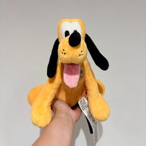 May include: A plush toy of Pluto, a yellow dog with black ears and a black nose. The toy has a pink tongue sticking out and is sitting upright. The toy is being held by a person.