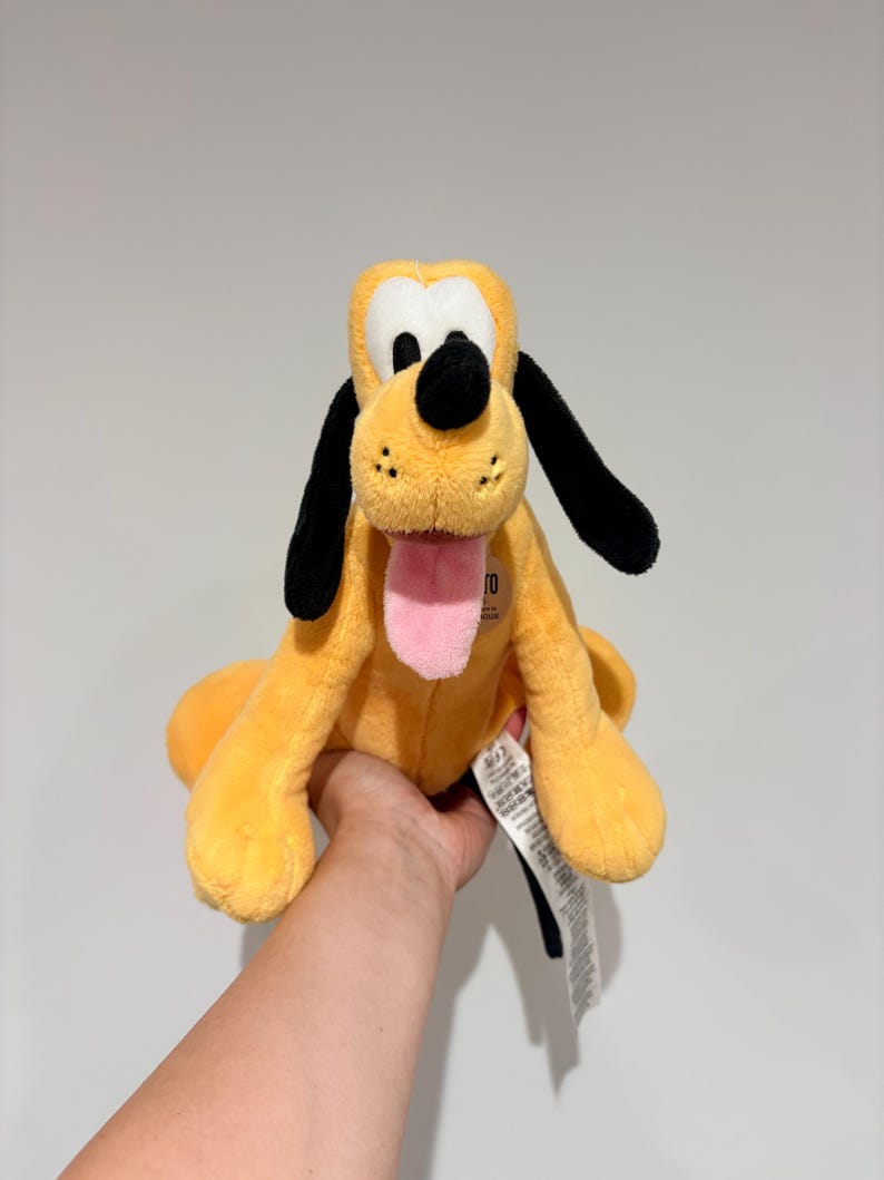 May include: A plush toy of Pluto, a yellow dog with black ears and a black nose. The toy has a pink tongue sticking out and is sitting upright. The toy is being held by a person.