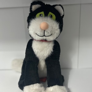 May include: A plush black and white cat with green eyes and a pink nose. The stuffed animal has a red collar and is sitting upright. The cat's fur is soft and fluffy, and it has a friendly expression.