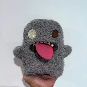 Op de afbeelding: Een grijs pluche monster speelgoed met knoopogen en een roze tong. Het speelgoed heeft een harige textuur en een brede, tandige grijns. Het monster heeft kleine armen en wordt door een hand omhoog gehouden.