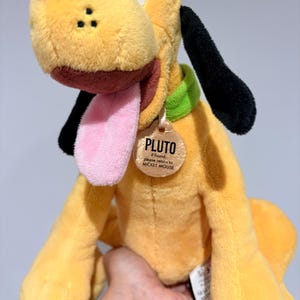 May include: A plush Pluto dog toy, featuring yellow fur, black ears, and a pink tongue. The toy has a green collar and a tag that reads "PLUTO". The toy is held in a person's hand.