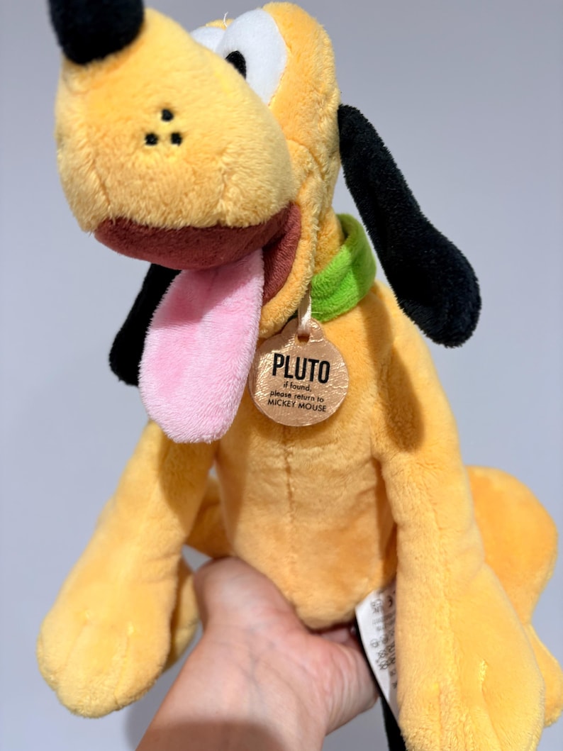 May include: A plush Pluto dog toy, featuring yellow fur, black ears, and a pink tongue. The toy has a green collar and a tag that reads "PLUTO". The toy is held in a person's hand.