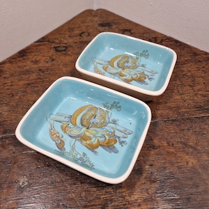 May include: Two small, square ceramic dishes with a light blue glaze. Each dish features a hand-painted design of a cluster of yellow and orange mushrooms on a green stem with green leaves.