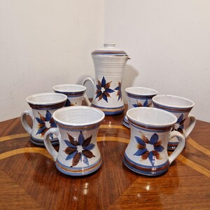 May include: A set of six white ceramic mugs with blue and brown floral designs and a matching jug. The mugs have handles and the jug has a lid.