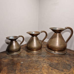 May include: A set of three graduated copper jugs with wide, flared rims, rounded bodies, and curved handles. The jugs feature a metallic finish with visible variations in tone and texture, arranged in a row from smallest to largest.