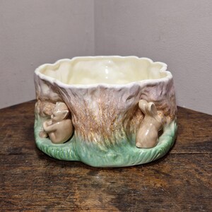 May include: A ceramic planter shaped like a tree stump with a scalloped rim. The exterior features a textured bark finish in shades of brown and pink, with a green base. A small sculpted figure of a seated elf and a rabbit are attached to the side.
