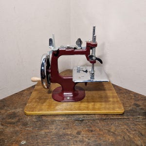 May include: A vintage-style manual sewing machine featuring a deep maroon metal frame, a polished chrome handwheel with a light-coloured wooden handle, and a silver-toned metal needle plate, all mounted securely onto a rectangular wooden base.