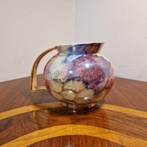 May include: A round ceramic jug with a gold handle. The jug is decorated with a colourful abstract design in shades of purple, pink, yellow, and brown.