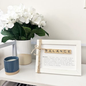 May include: White framed print with the word "BALANCE" spelled out in Scrabble tiles. The definition of balance is written below the tiles.