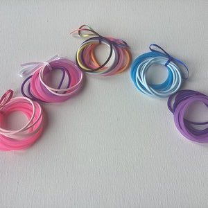 May include: Assortment of colorful silicone hair ties in pink, purple, blue, and rainbow colors. The hair ties are arranged in a semi-circle on a white background.