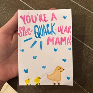 May include: A handwritten card with the text "You're a Spec-Quack-ular Mama!" in pink and blue lettering. The card features a drawing of three yellow ducklings with blue hearts scattered around them.