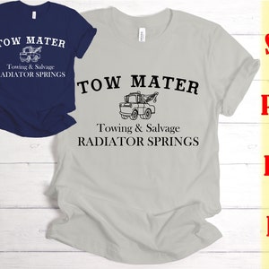 Puede incluir: Una camiseta gris con el texto "Tow Mater" y una imagen de dibujos animados de una grúa. El texto "Towing & Salvage" y "Radiator Springs" también está impreso en la camiseta.