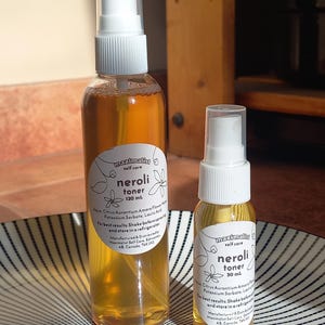 May include: Two glass bottles of neroli toner, one 120 ml and one 30 ml, with white labels and black text. The bottles are on a black and white striped surface.