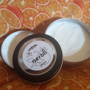May include: A small, black tin container with a clear lid. The lid has a white label with the text "maximalist self care" and "neroli lotion". The tin is filled with a white lotion.