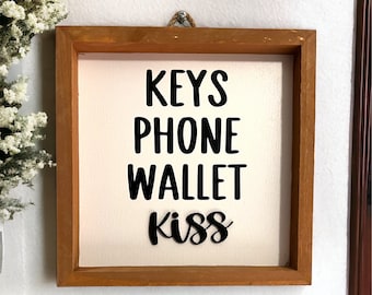 May include: A wooden framed sign with the words "KEYS PHONE WALLET kiss" in black lettering on a white background.