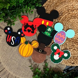 May include: A set of eight felt Mickey Mouse head ornaments, each decorated with a different theme. The ornaments include a pumpkin, a turkey, a Christmas wreath, a leprechaun, an Easter egg, a heart, a USA flag, and a classic Mickey Mouse.