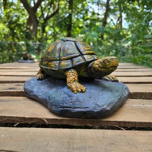 May include: A sculpted turtle figurine with a textured, multi-colored shell in shades of green, yellow, and brown. The turtle is positioned on a dark, flat, rock-like base, featuring detailed scales on its head and legs, with a forward-facing gaze.
